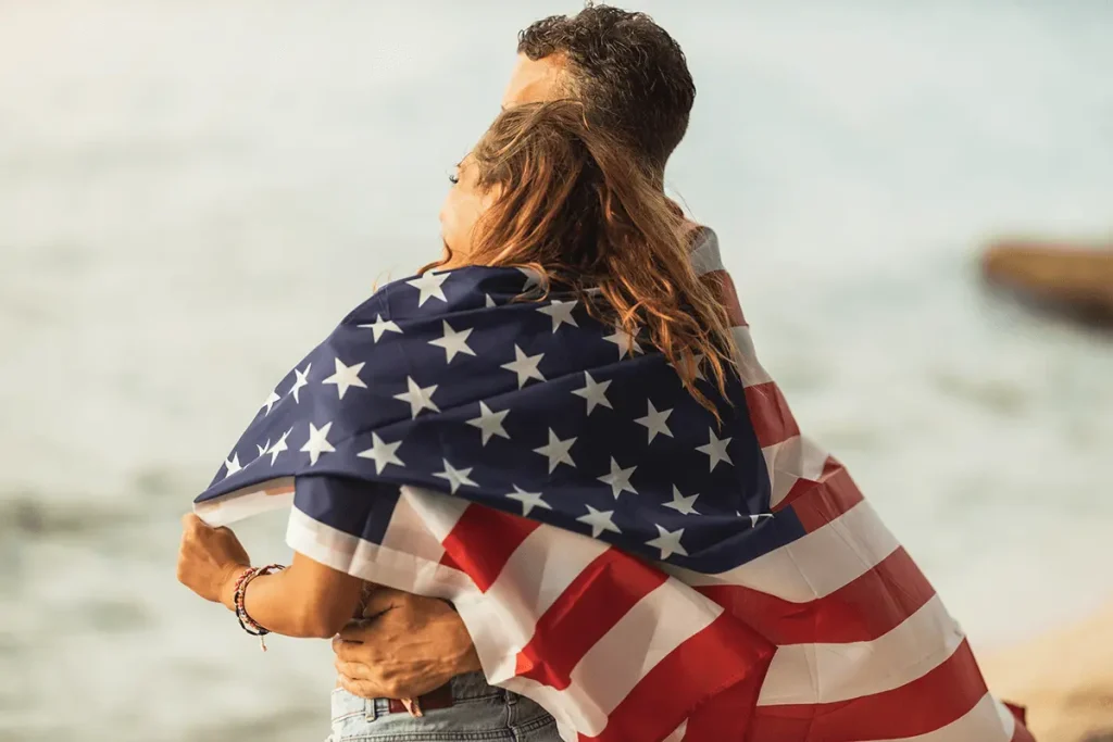 Engaged couple cuddling wrapped in American flag on the beach, symbolizing K-1 visa journey