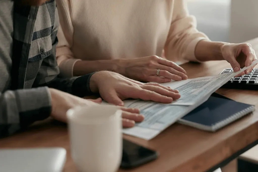 Couple sitting at a table at home, closely reviewing financial documents with a keyboard, notebook, and coffee mug nearby.
