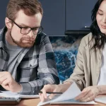 Interracial couple sitting at a wooden table at home, reviewing immigration paperwork together with a laptop, calculator, and documents spread out in front of them.