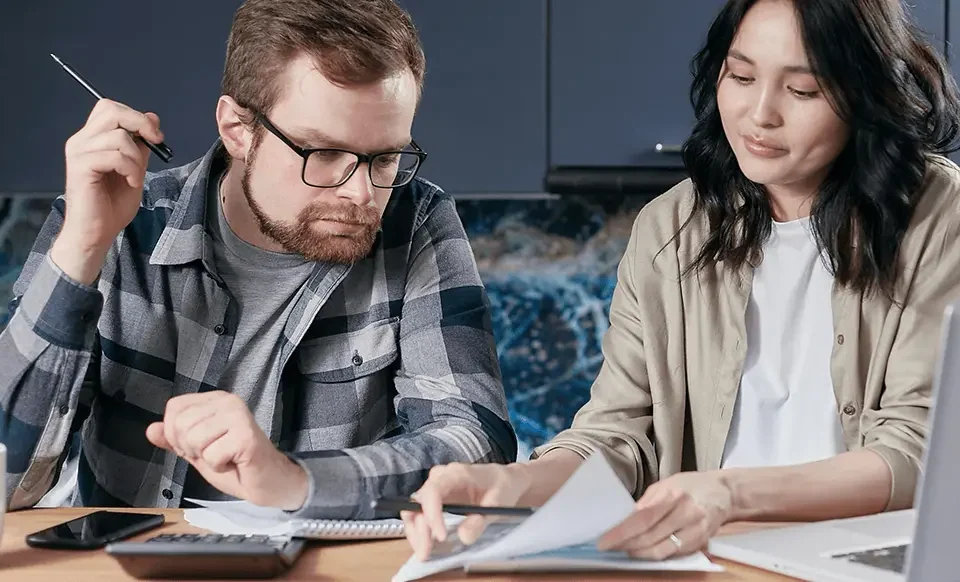 Interracial couple sitting at a wooden table at home, reviewing immigration paperwork together with a laptop, calculator, and documents spread out in front of them.