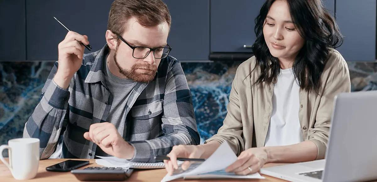 Interracial couple sitting at a wooden table at home, reviewing immigration paperwork together with a laptop, calculator, and documents spread out in front of them.