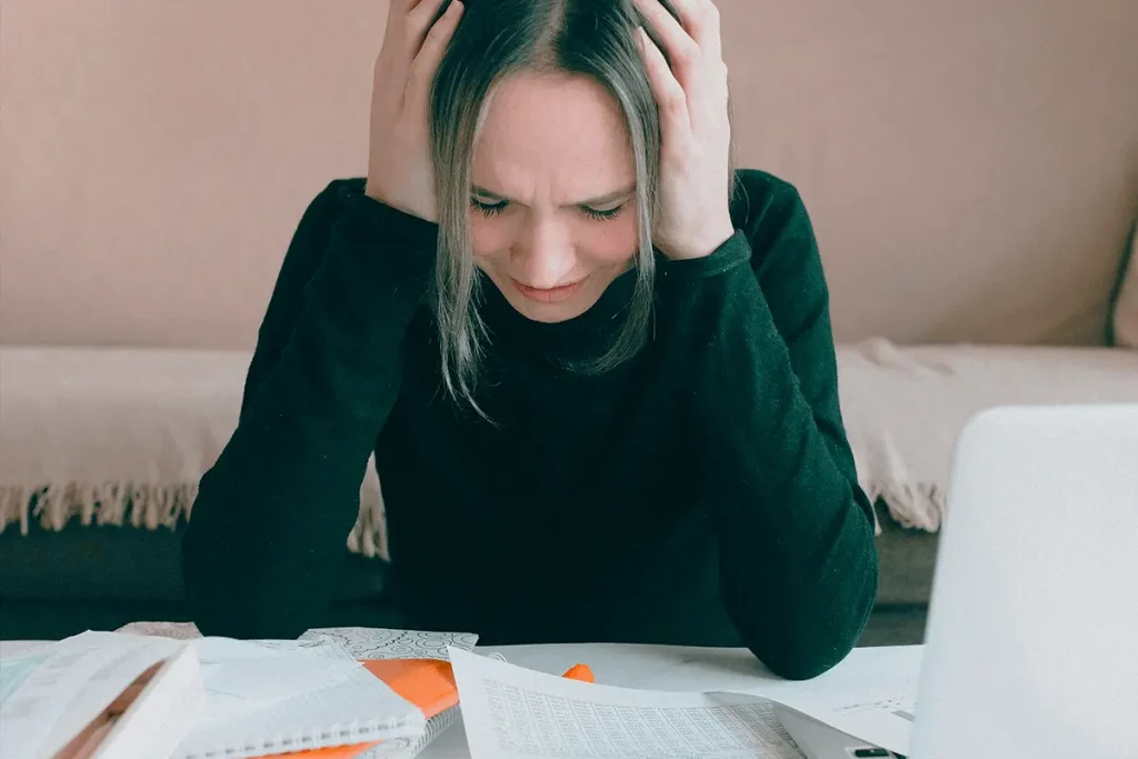 Stressed woman sitting at a table at home, resting her head on one hand while surrounded by scattered paperwork, a laptop, and a calculator.
