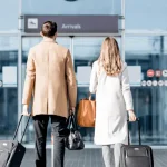 A couple walking into an airport arrivals entrance, each pulling a suitcase and carrying bags, symbolizing international travel and relocation.