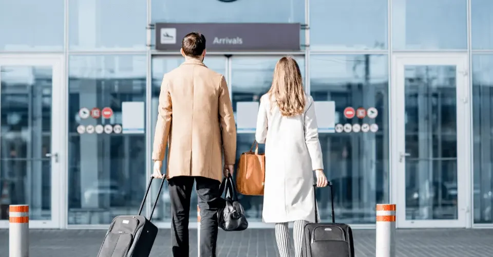 A couple walking into an airport arrivals entrance, each pulling a suitcase and carrying bags, symbolizing international travel and relocation.