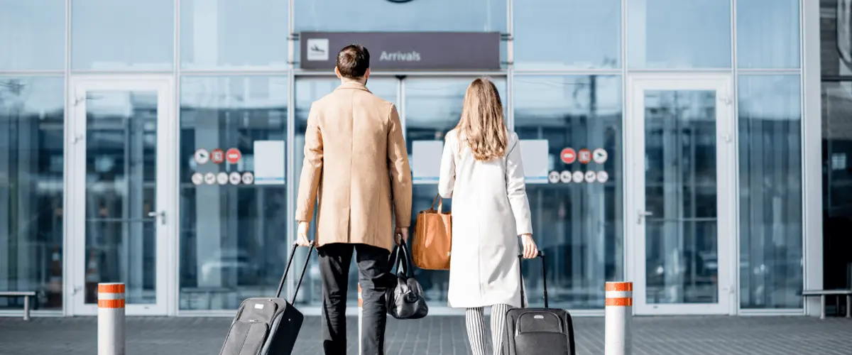 A couple walking into an airport arrivals entrance, each pulling a suitcase and carrying bags, symbolizing international travel and relocation.