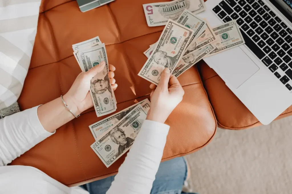 A person counting U.S. dollar bills beside an open laptop, representing financial planning and expenses.