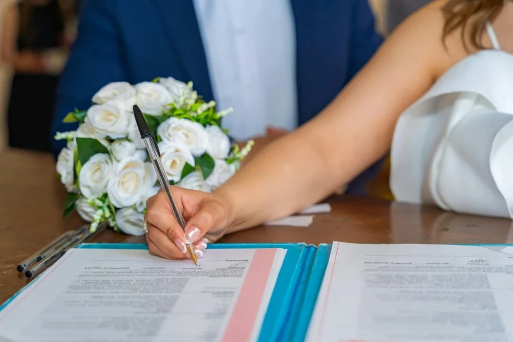 Bride in white dress signing marriage certificate with pen at wedding ceremony with bouquet