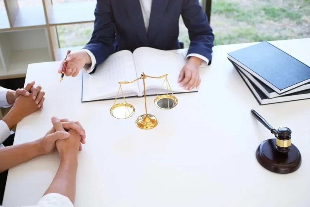 Lawyer in a suit meets with two clients at a desk, with an open notebook, golden scales of justice, and a gavel visible.