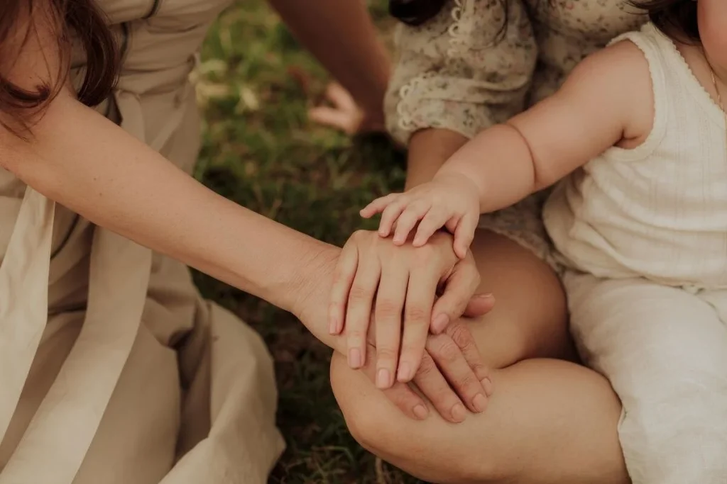 Close-up of a child’s hand resting on top of two adults’ hands as they sit together on grass, warm and intimate family moment.