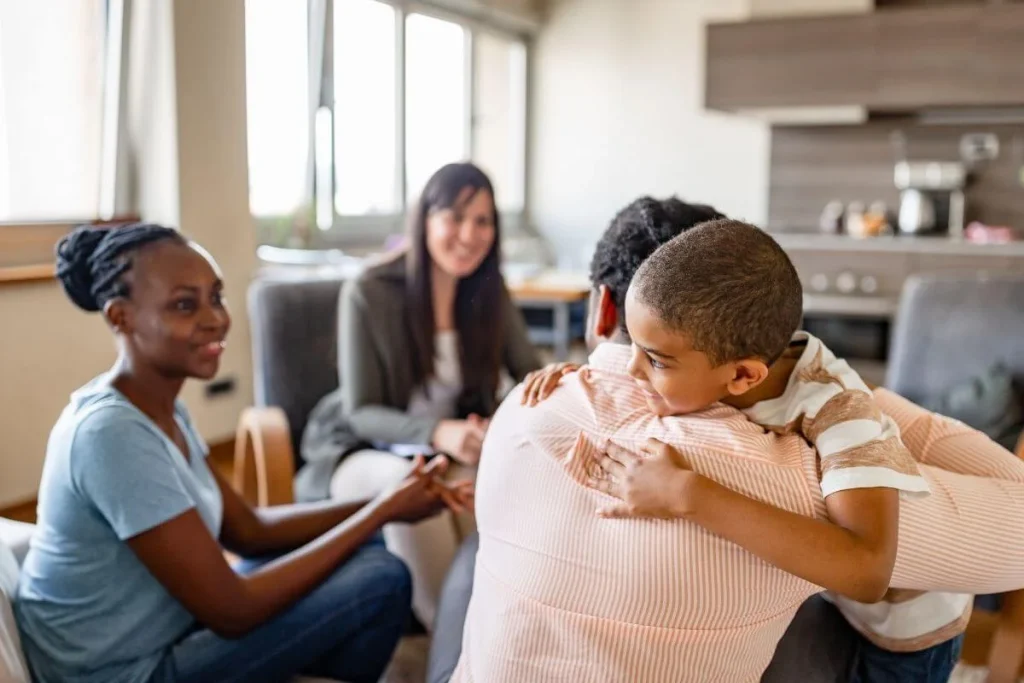 Smiling child hugs an adult in a cozy living room while another adult and a counselor look on, capturing a joyful family reunion moment.