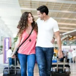 Smiling couple walk arm-in-arm through a bright airport terminal, each pulling a rolling suitcase.