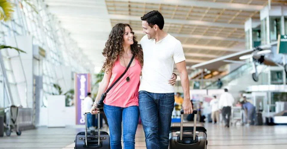 Smiling couple walk arm-in-arm through a bright airport terminal, each pulling a rolling suitcase.