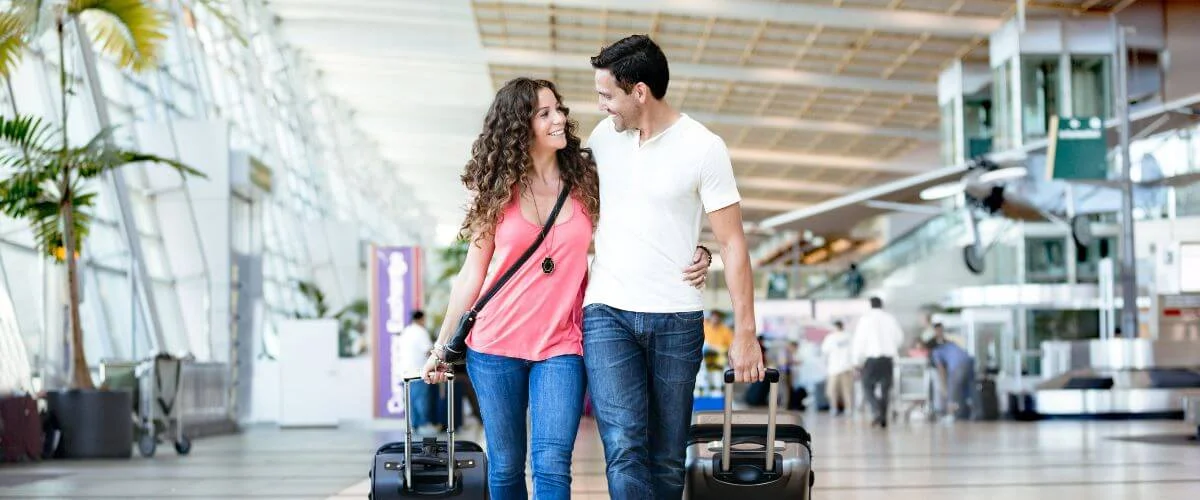 Smiling couple walk arm-in-arm through a bright airport terminal, each pulling a rolling suitcase.